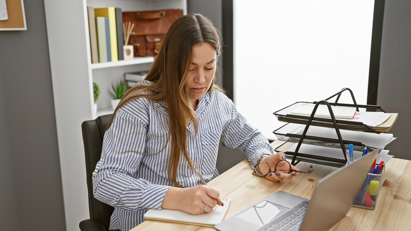 Focused woman writing notes in a modern office setting, with laptop and organized documents
