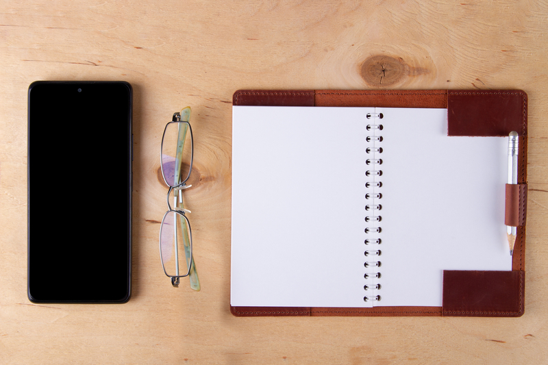 Eyeglasses digital phone with blank black screen and empty paper pad lie on wooden desk