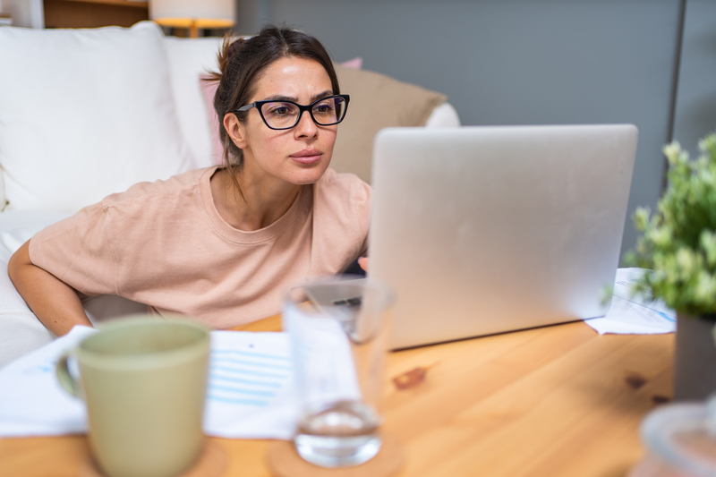 Female freelancer reviewing tax documents on laptop at home coffee table with papers and coffee nearby.