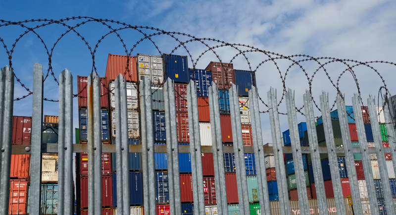 Shipping containers stored behind razor wire representing supply chains rerouting after U.S. tariffs enacted